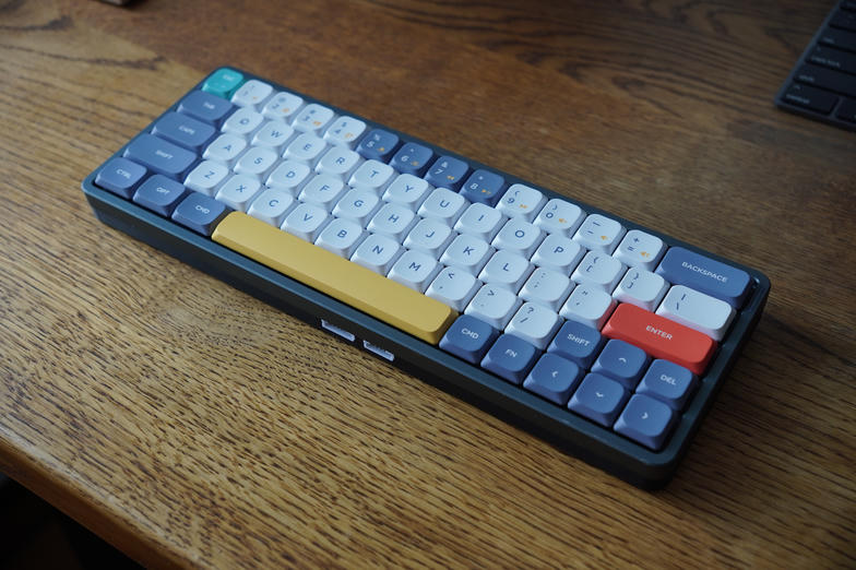 a NuPhy Air60 on a wooden desk, with blue and white keycaps, a yellow spacebar and a red enter key, sitting on a black printed base